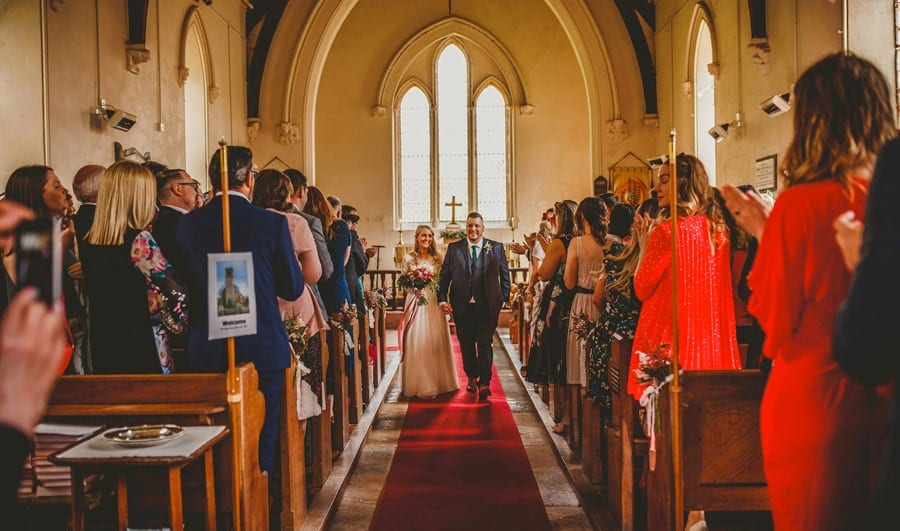 The bride and groom walk down the aisle of the Church together as wedding guests stand up and clap their hands