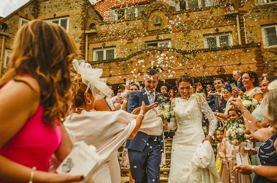The wedding party throw confetti as the bride and groom walk down the stone steps at the front of Voewood