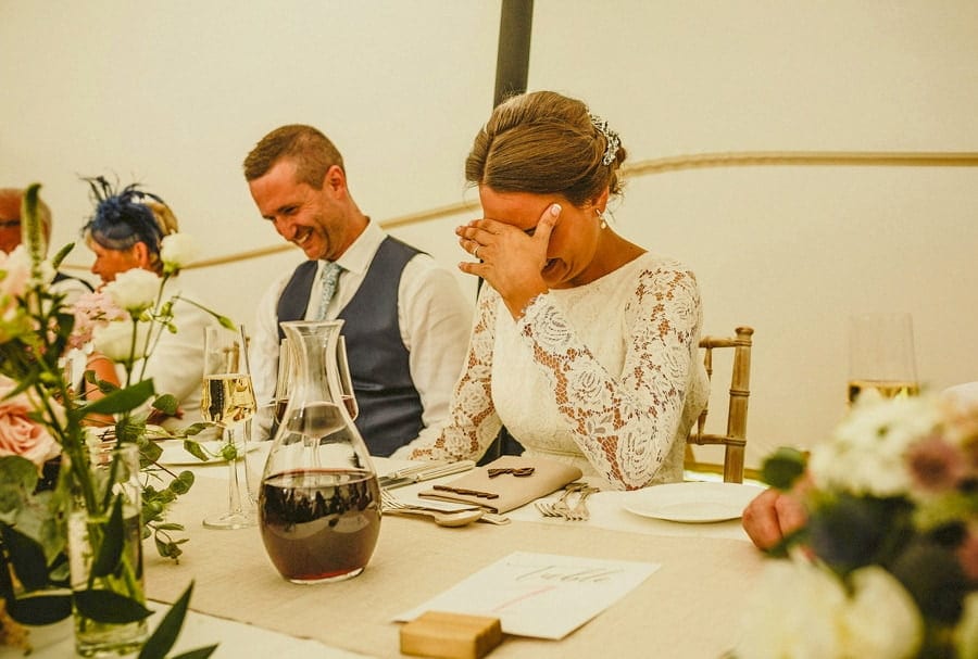 The bride puts her hands over face and laughs at one of the speeches in the marquee at Voewood House in Norfolk