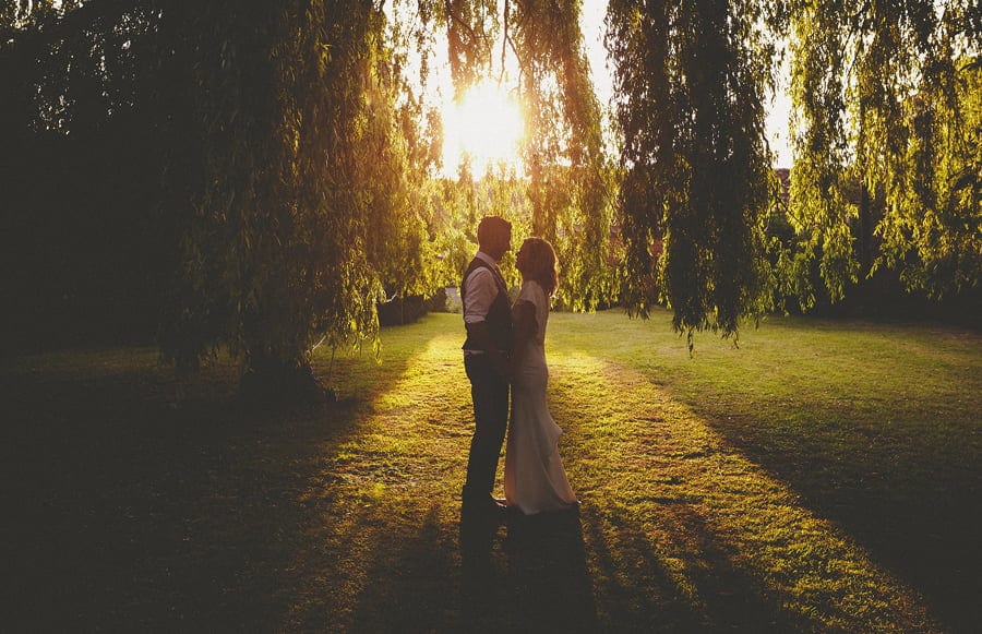 The bride and groom embrace each other under a weeping willow tree as the sun sets