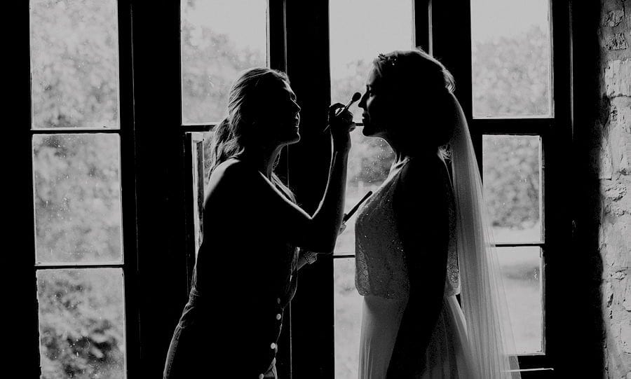 The makeup artist brushes foundation onto the bride's face next to a large window in the old barn at Childerley