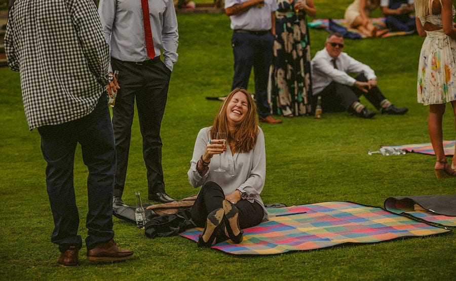 A lady lies on a multi coloured blanket holding a glass of wine and laughs with friends