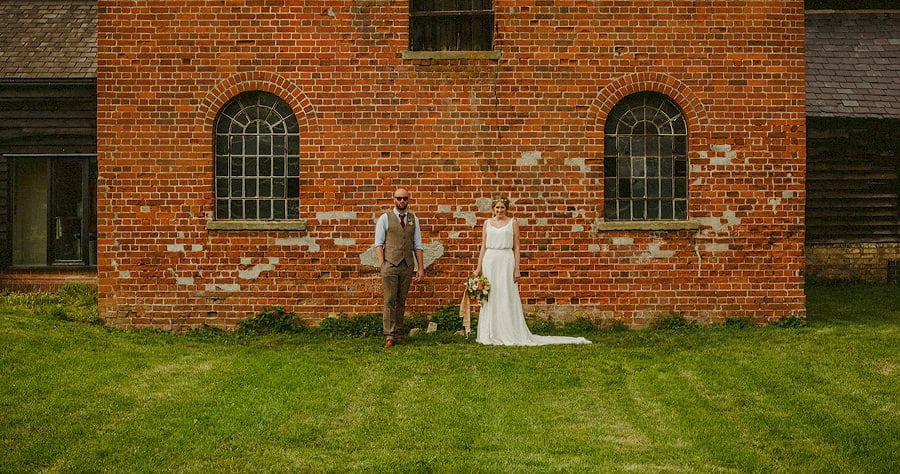 The bride and groom stand next to a large wall and pose for a photograph at Childerley Estate