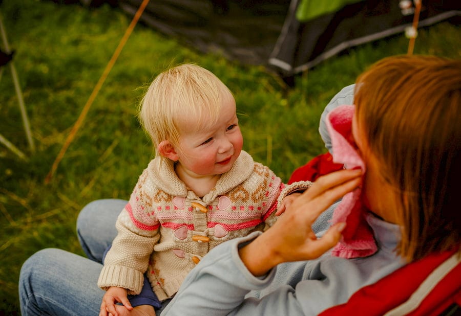 A little girl sits on her mother's lap and watches her mother put a red blanket across her face