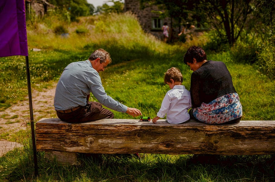 A little boy sits on an old tree stump with his grandparents
