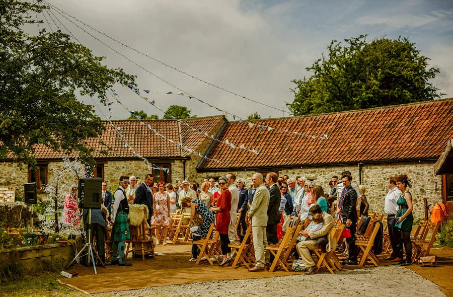 The guests stand and wait for the bride and her father in the courtyard at Fernhill Farm