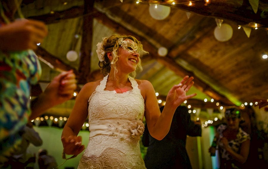 The bride dancing and wearing a gold mask smiles to a friend on the dancefloor