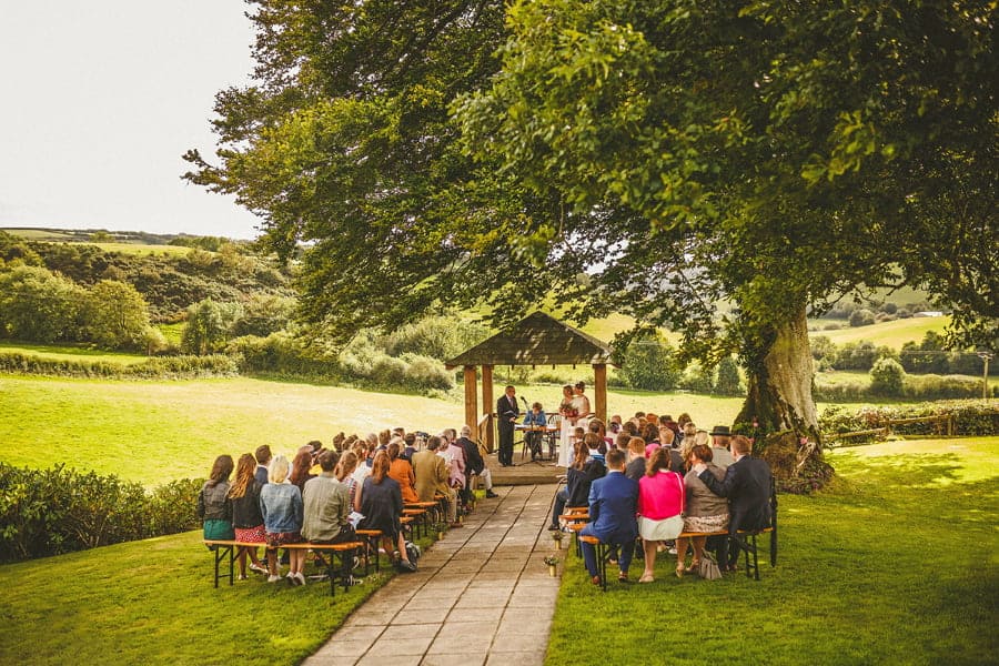 An outdoor wedding ceremony with wedding guests sat on long wooden benches in front of the bride and groom in the countryside