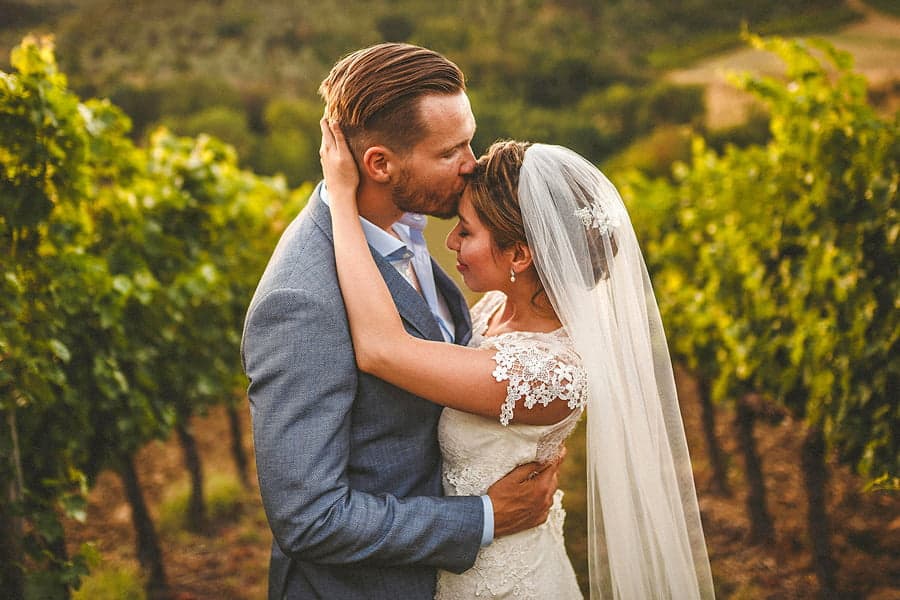 The groom holds the bride and kisses her on the forehead in a field as she puts both her hands on the back of his head