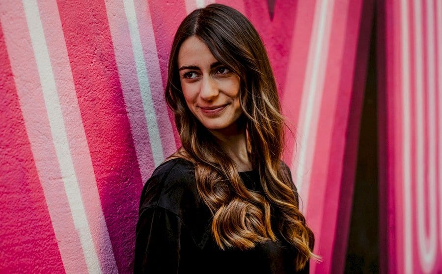 A lady with long brunette hair looks away from the camera and poses for a photograph next to a wall with red painted lines across it