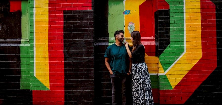 A man and lady stand against a multi coloured painted wall and look at each other