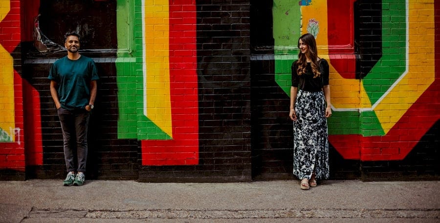 A man and lady stand apart from each other and pose for a photograph against a multi coloured painted wall