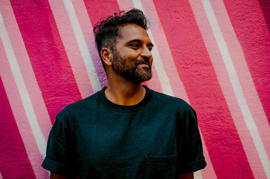 A man looks away and stands with his back against a large wall painted in red, white and pink
