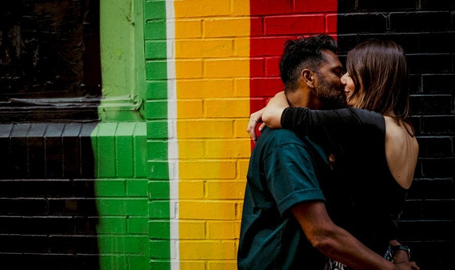 The couple embrace each other and kiss against a large wall painted in red, yellow, green and black