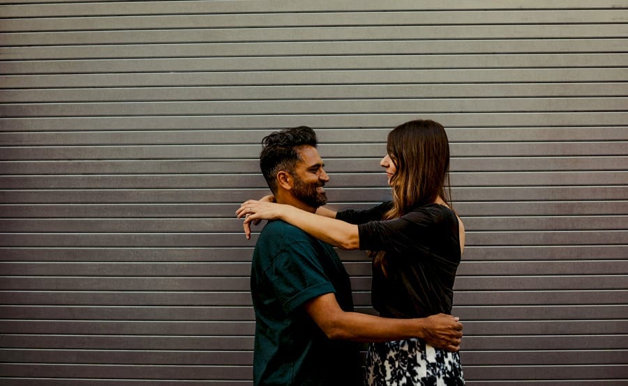 A man holds the lady around the waist and the lady places her arms across the man's shoulders as they look at each other against a large metal shutter