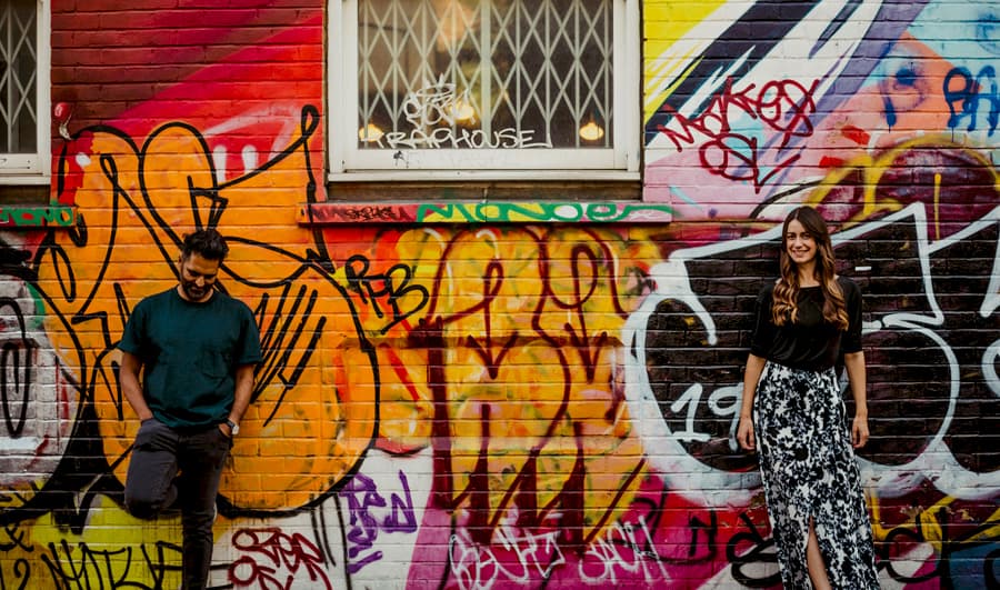 The man looks down on the floor and the lady looks at the camera as they pose for a photograph against a large brick multi coloured wall