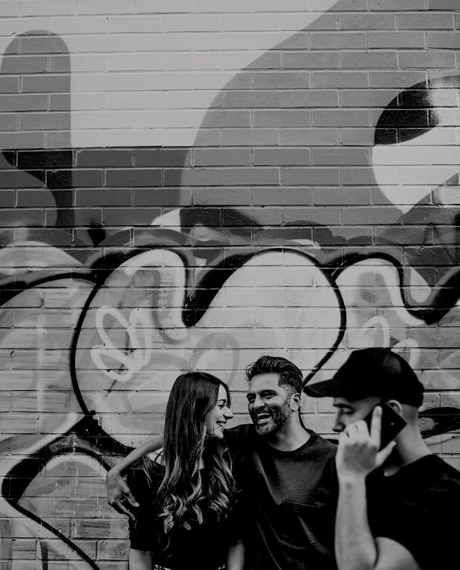 A man and lady share a joke with each other and laugh against a wall as a man with a mobile phone and a black cap walks passed