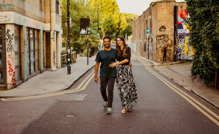 A lady and a man walk side by side and smile as they pose for a photograph in the middle of a road in Shoreditch in London