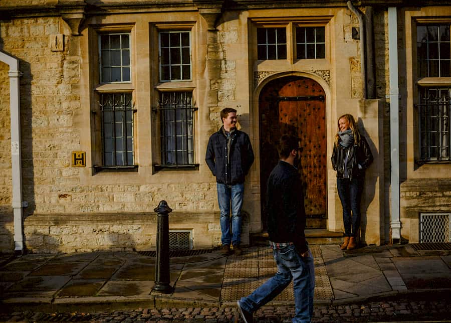 A man and lady stand next to each other and smile against a building as a man walks passed and looks at them in Oxford city centre