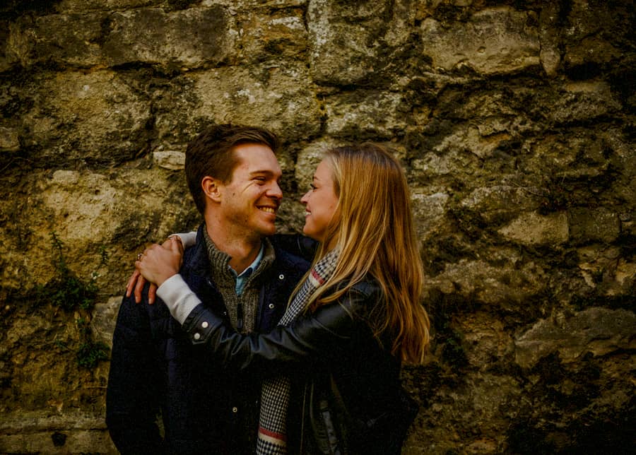 A lady places her arms around the shoulders of a man and they smile at each other against an old brick wall in Oxford