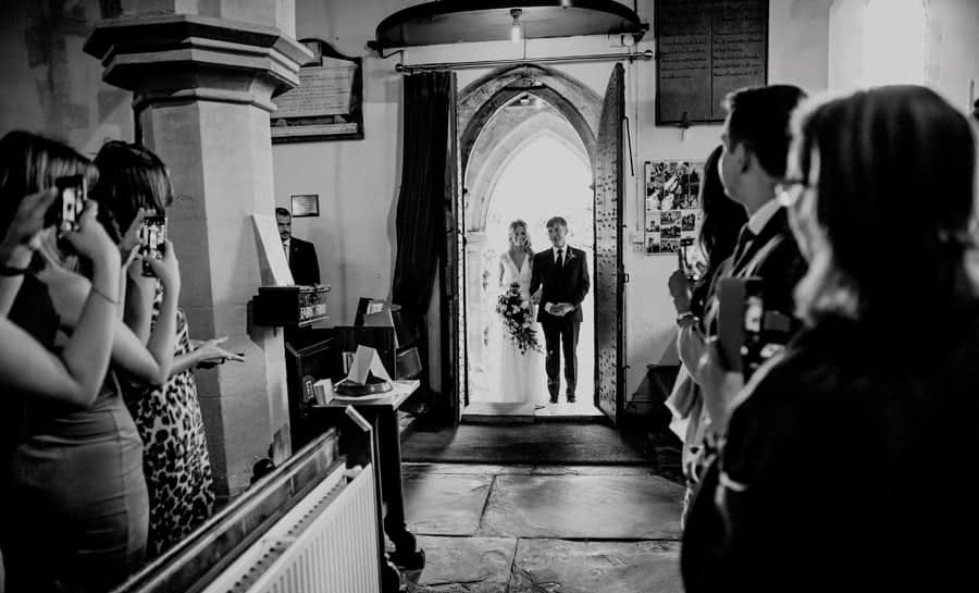 The bride and her father stand at the door of the Church as wedding guests stand and photograph them