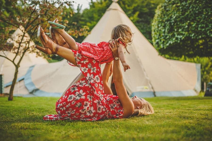 A lady lies on the grass in front of a tipi and lifts a little girl up in the air