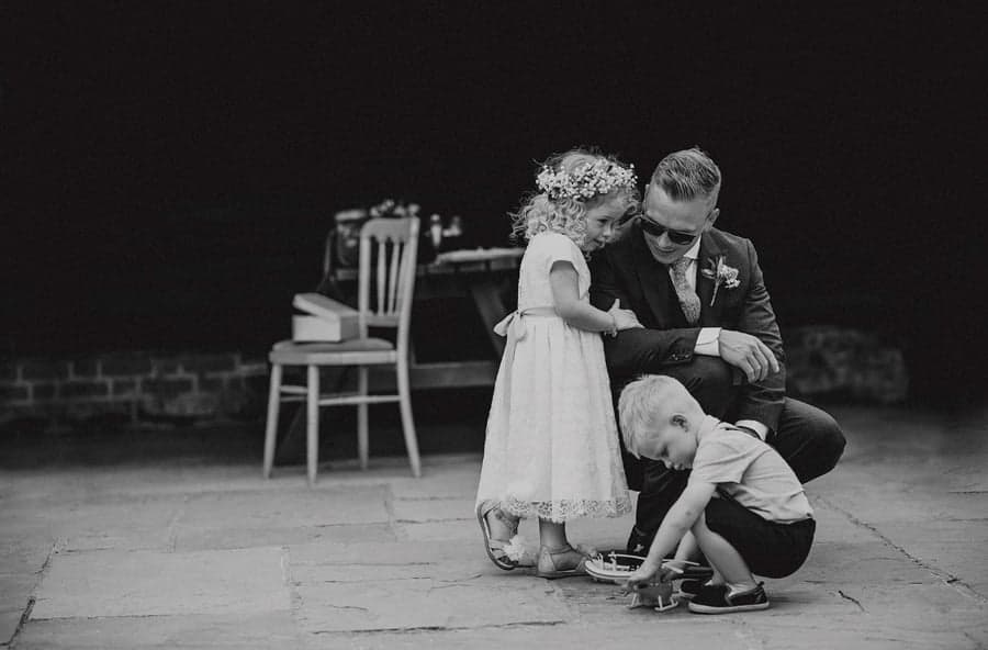 The groom kneels down and listens to his daughter in a courtyard with a little boy in front of them playing with a toy helicopter