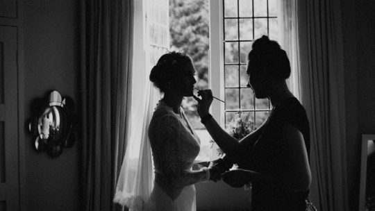 A makeup artist applies lipstick to the lips of the bride as she stands next to a large window in her room