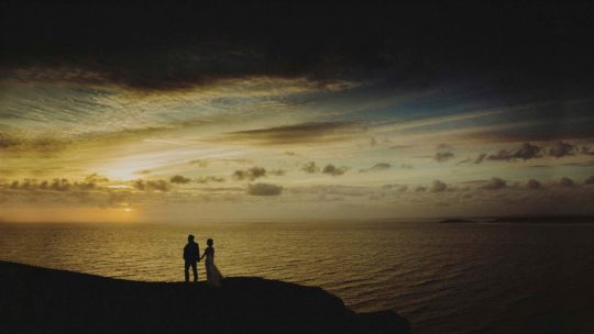 The bride and groom hold each others hand and stand next to each other on the edge of a cliff and watch the sun go down over the sea
