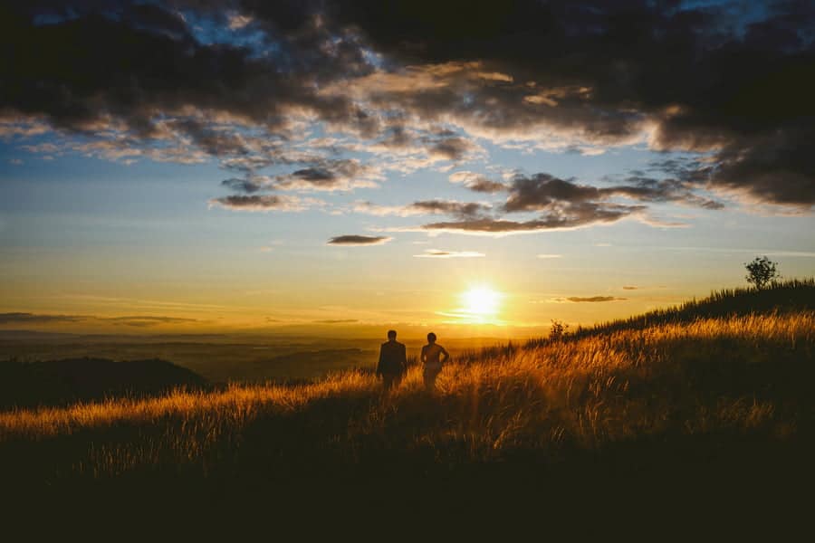 A bride and groom walk through grass next to each other on a hill as the sun goes down in the distance