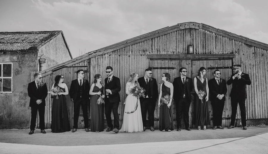 The bride and groom stand in front of large shed and talk with the wedding party