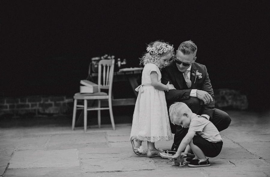 The groom kneels down and listens to his daughter in a courtyard with a little boy in front of them playing with a toy helicopter