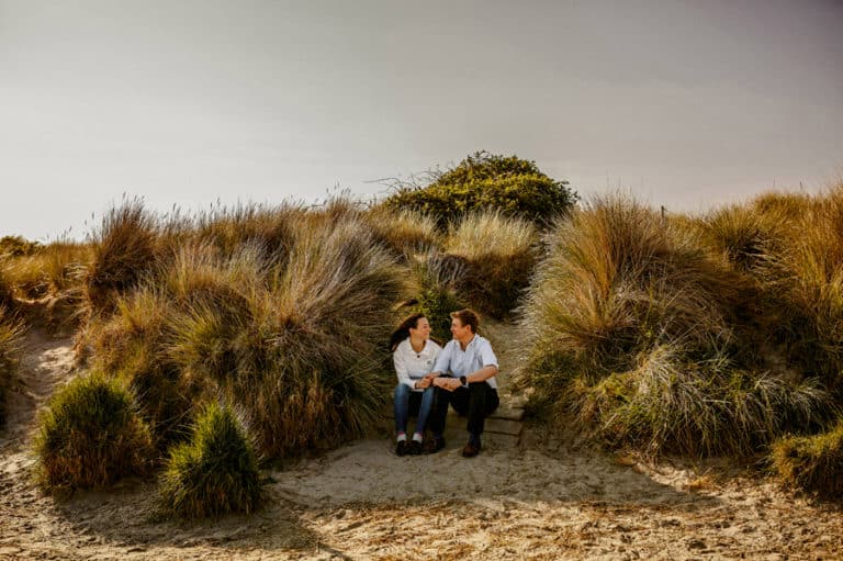A man and lady sit next to each other and smile at one another against a back drop of sand dunes