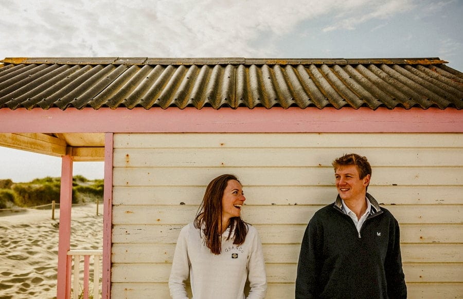 A man and lady stand at the side of a beach hut on the beach in West Wittering