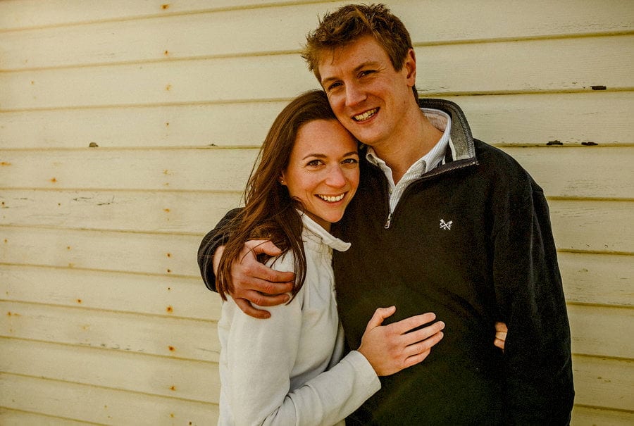 A lady and a man hold each other in their arms and pose for a photograph next to a beach hut on West Wittering beach