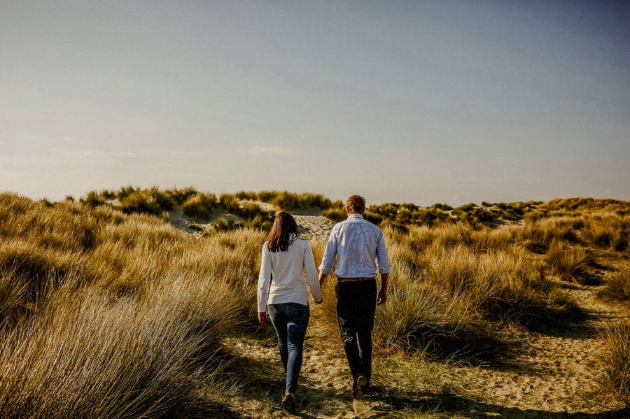 A man and lady hold each others hand and walk side by side along the sand dunes at West Wittering beach