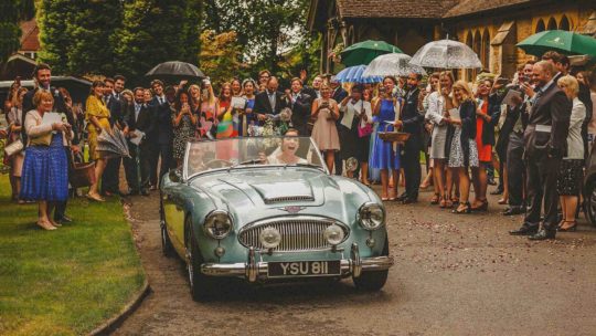 The bride and groom leave the church in a vintage car as the wedding party watch behind them