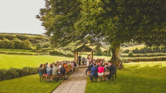 An outdoor wedding ceremony with wedding guests sat on long wooden benches in front of the bride and groom in the countryside